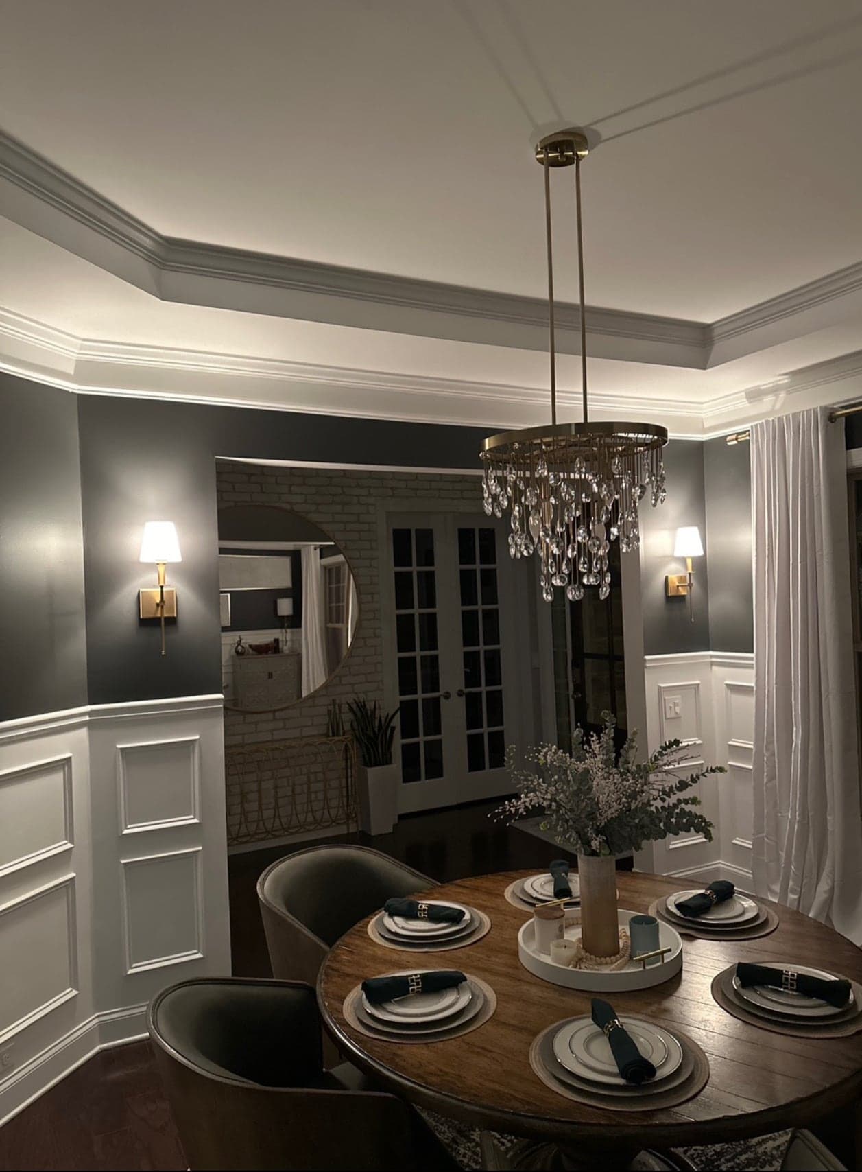 A dining room at night, a round crystal chandelier on long brass rods over a set table and a brass wall sconce with a white shade lit on each side wall