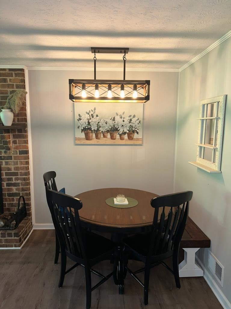 A long black-framed light with five bare bulbs, lit, hanging over a round wooden table and black chairs, a brick fireplace to one side