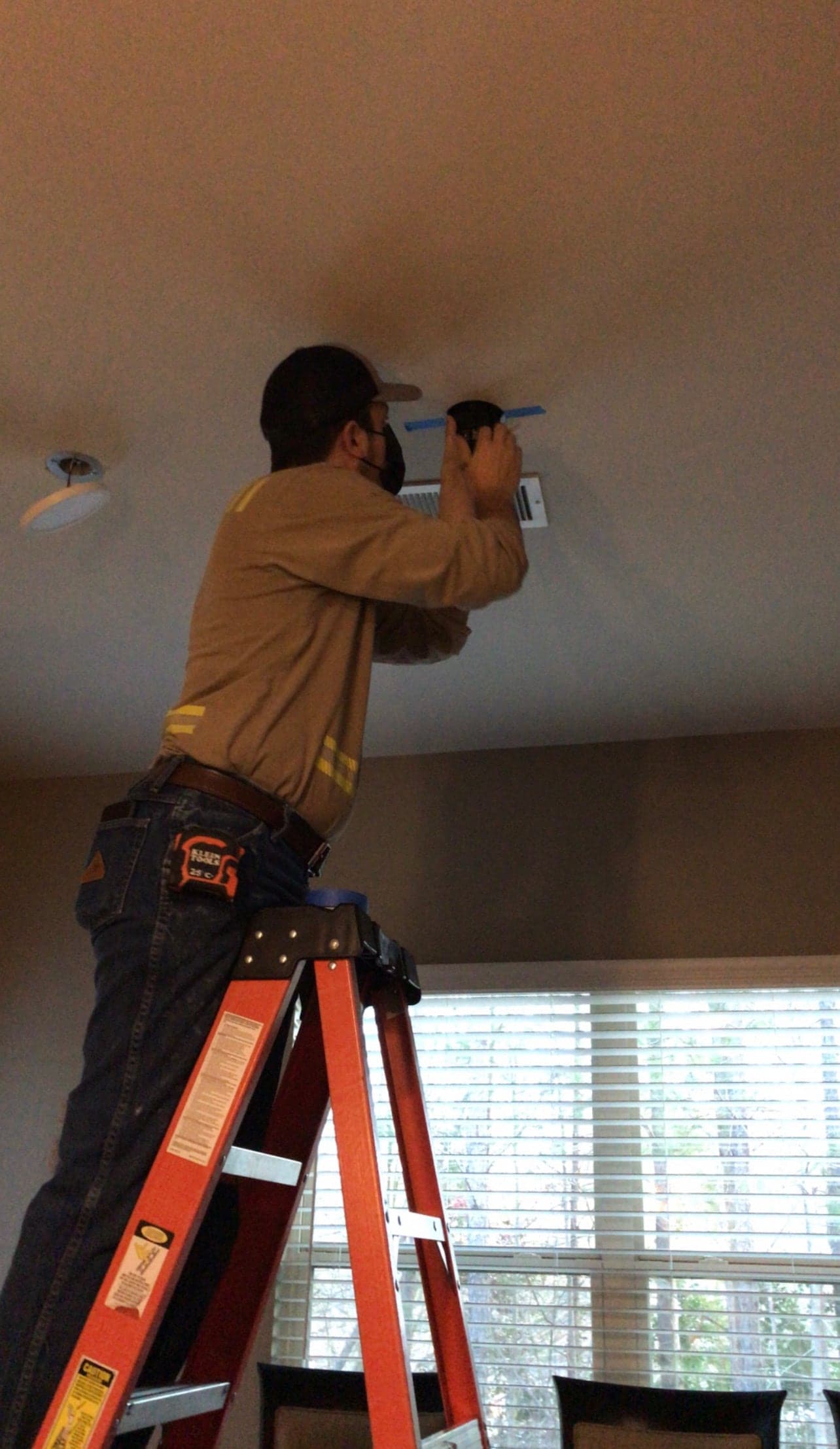 An electrician on an orange stepladder working at a ceiling marked out with blue tape, a slim recessed light hanging from its hole a few feet away