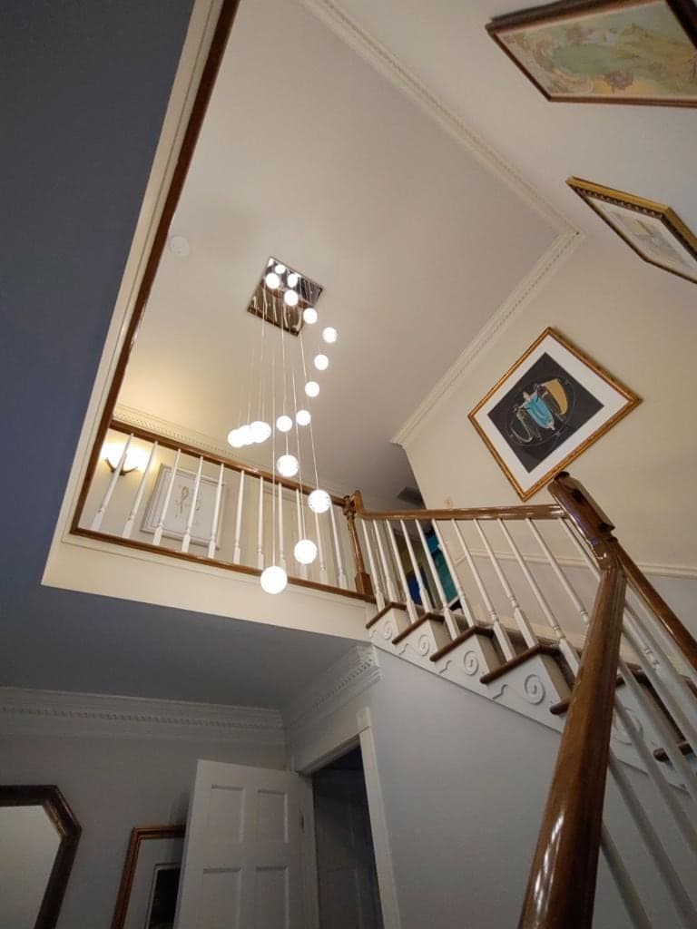 Looking up a two-story stairway at a cascade of glowing glass globes hanging on long cords from a square ceiling plate beside the upstairs landing